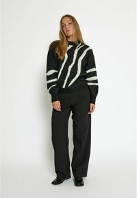 Großer Strickpullover in Schwarz mit weißen abstrakten Streifen, mit gerippten Bündchen und Saum, kombiniert mit weit geschnittenen schwarzen Hosen und lackierten Schuhen.