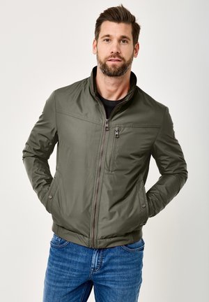 Man with brown hair and beard wearing a zipped green jacket and blue jeans, standing with hands in pockets against a plain background.