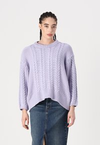 Pull en maille lavande avec un motif de torsades épaisses, ourlet côtelé et manches larges, associé à une jupe en denim avec une fente à l'avant.
