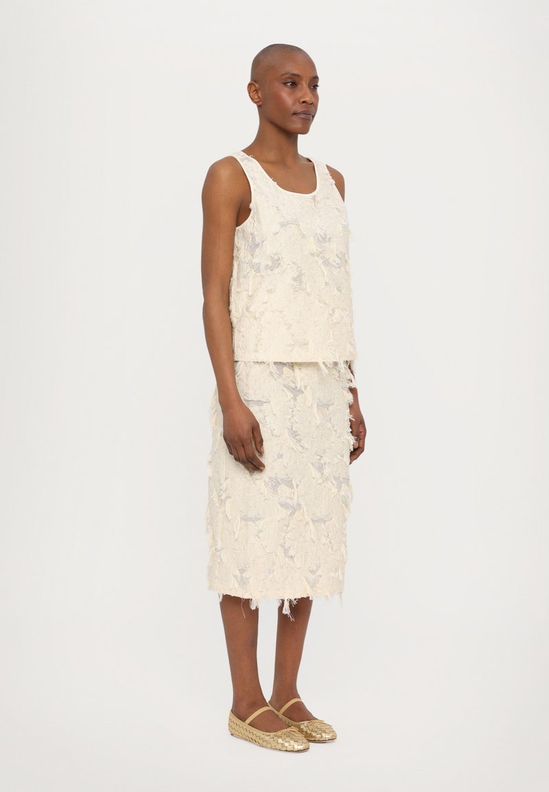 Linnen twee-delig outfit in crème met een getextureerd bloemenpatroon, bestaande uit een mouwloos topje en een rok tot op de knie, gecombineerd met gouden instapschoenen.