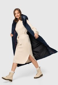 Langer, dunkelblauer Pufferwesten mit Kapuze, geöffnet, um ein dunkles Innenfutter zu zeigen. Beige gestricktes Kleid mit Taillengürtel und seitlichem Schlitz. Beige Schnürstiefel.