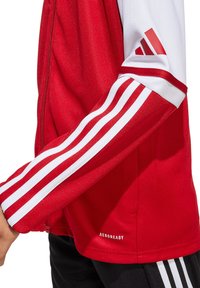 Rote Sportjacke aus strukturiertem Stoff mit weißen drei Streifen als Akzent an den Ärmeln. Mit "AEROREADY"-Label nahe dem Saum. Mit Reißverschluss vorne.