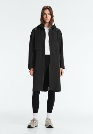 Vrouw staand, gekleed in een zwarte lange jas over een wit shirt, zwarte legging, witte sokken en beige sneakers op een effen witte achtergrond.