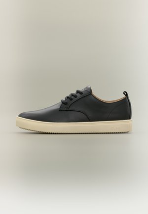 Zwarte lage leren sneaker met crèmekleurige rubberen zool en zwarte veters tegen een effen beige achtergrond.