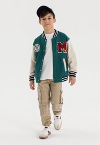 Tealfärgad och crèmefärgad varsityjacka med bokstaven "M", broderad patch, vit t-shirt, beige cargo-byxor och vita sneakers med detaljer.