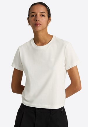 Młoda kobieta z krótkimi włosami, nosząca małe kolczyki koła, prosty biały t-shirt z krótkim rękawem i czarne spodnie, stojąca z rękami za plecami.