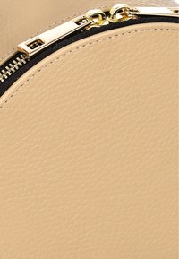 El bolso circular beige presenta un material sintético texturizado, un cierre de cremallera en tono dorado y un ribete negro a lo largo del borde de la cremallera.