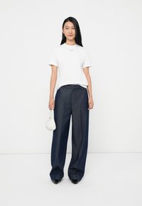 Fiatal nő, hosszú fekete hajjal, fehér Calvin Klein pólót viselve, széles szárú sötét farmernadrággal, fekete hegyes orrú cipővel, és egy kis fehér kézitáskával a kezében.