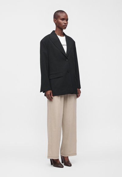 Femme debout portant un blazer noir oversize, une chemise blanche, un pantalon beige large et des chaussures à talons marron sur un fond uni.