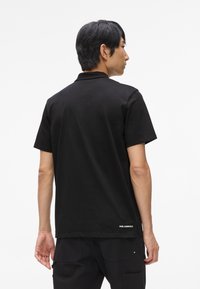 Čierna polo košeľa s krátkym rukávom vyrobená z bavlny, s golierom a vyšitým logom na zadnej strane pásu. Hladká textúra, klasický strih.