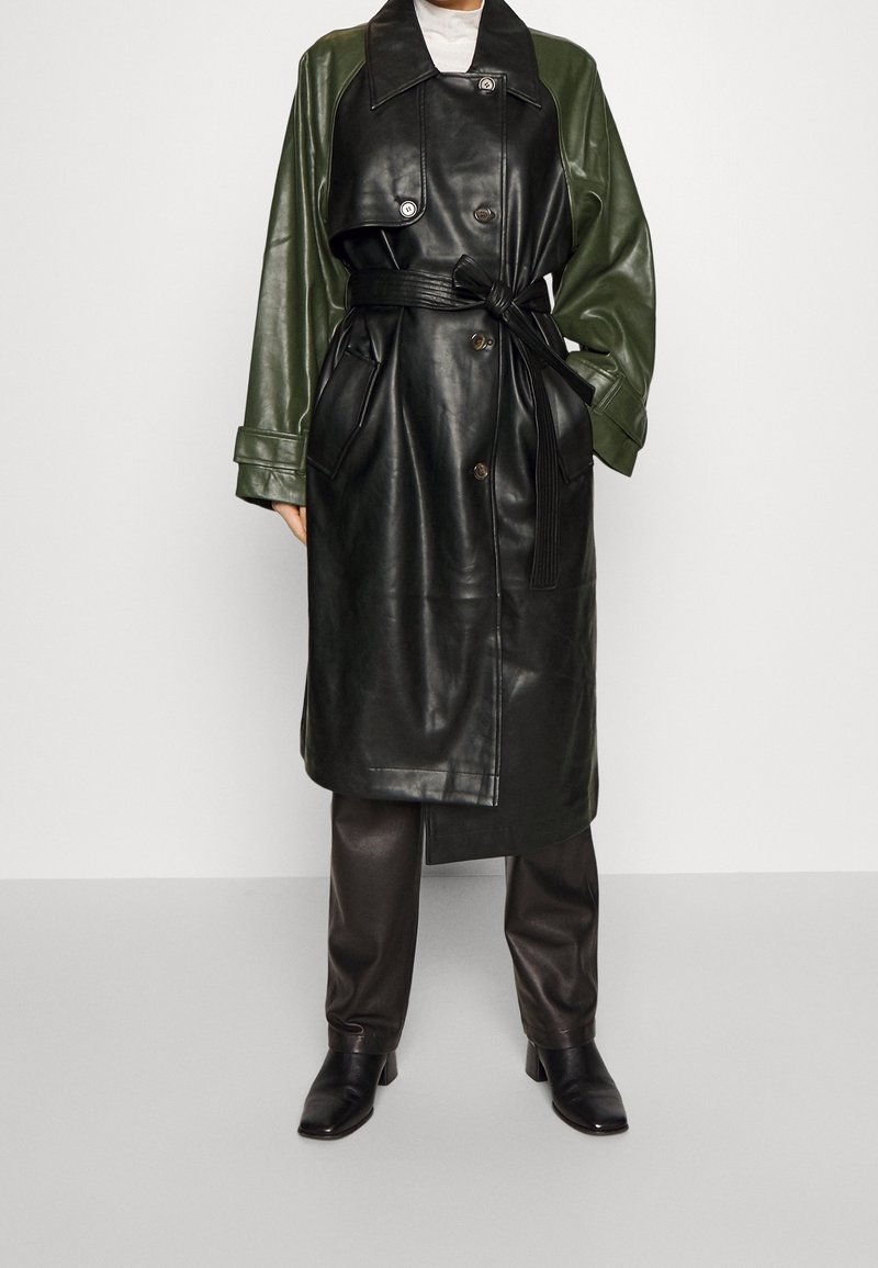 Černý a tmavě zelený kožený trench coat s opaskem kolem pasu, velkým límcem a knoflíkovým zapínáním. Má dlouhé rukávy a hladkou texturu.
