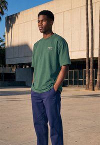 Jeune homme debout à l'extérieur, les mains dans les poches, portant un t-shirt vert et un pantalon bleu, près d'un bâtiment moderne en béton et des palmiers.