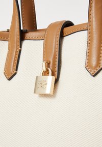 Beige canvas tote bag with brown leather straps and accents. Gold padlock detail attached to the strap. Textured surface and sleek design.