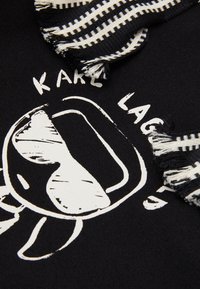 Černá látka s bílým náčrtem obličeje nosícího sluneční brýle, částečně zakrytá černobílým pruhovaným třásňovým šátkem. Částečně viditelný text.