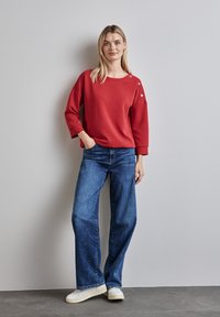 Röd långärmad sweatshirt med detaljer med knappar på sidan, parat med vida blå jeans och vita sneakers på en grå bakgrund.