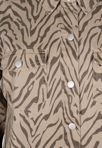 Tan denim shirt met zwarte zebra-strepen, voorzien van zilveren drukknoopjes en borstzakken, die de contrasterende patronen en texturen benadrukt.
