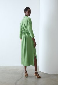 Lang groene jurk van een textuurstof, met een elastische taille, driekwart mouwen en een zijsplit. Gemaakt met beige hakken.