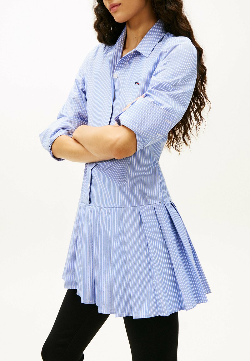 Donna con capelli lunghi e ricci che indossa un vestito camicia a righe sottili blu e bianche con maniche risvoltate e leggings neri, braccia incrociate.