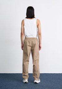 Beige elastic-waist pants made of lightweight fabric, paired with a white sleeveless top. Model shown from the back on a blue carpet.
