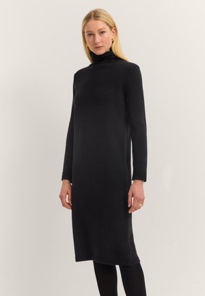 Blonde Frau, die ein schwarzes Langarm-Turtleneck-Strickkleid mit schwarzen Strumpfhosen trägt und vor einem schlichten, hellen Hintergrund steht.