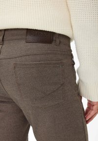 Braun strukturierte Hosen mit einem sichtbaren Markenetikett auf der Rückseite, mit einem taillierten Design und Taschen. Der Ärmel eines Pullovers ist teilweise sichtbar.