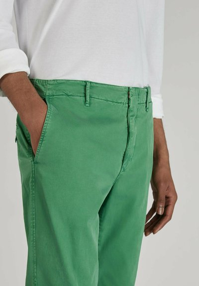 Chinos de algodón verde con una textura suave, presentando una cremallera, lazos para cinturón y bolsillos laterales. Combinados con una camisa blanca de manga larga.