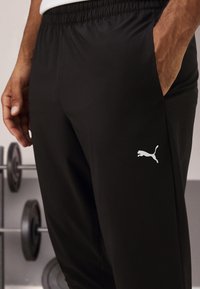 Zwarte sportbroek van gladde stof, met een elastische tailleband, zijzakken en een klein wit Puma-logo op de dij.