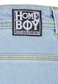 Svetlomodrá denimová tkanina má čiernu nálepku s bielym textom "HOME BOY loud-couture", umiestnenou na páse. Žlté švy sú zvýraznené.