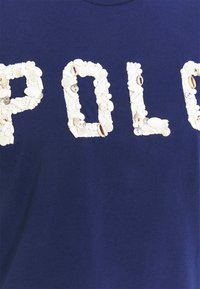 Marineblauwe stof met de letters "P," "O," en "L" gevormd uit verschillende witte schelpen en kleine kralen.