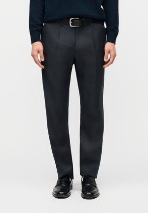 Férfi fekete öltönynadrágban, fekete övvel, tengerészkék pulóverben és fekete bőrcipőben, egyszínű háttér előtt állva.