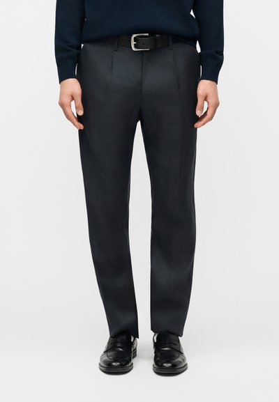 Homme portant un pantalon de costume noir avec ceinture noire, un pull bleu marine et des chaussures en cuir noir, debout devant un fond uni.