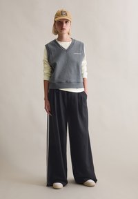 Grauer ärmelloser Pullover mit V-Ausschnitt, darüber ein cremefarbenes Langarmshirt. Lockere schwarze Hose mit weißen Seitenstreifen und beigefarbene Schuhe.