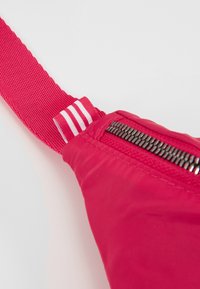 Röd tygpåse med en strukturerad yta, utrustad med en svart dragkedja och ett rosa band med vita ränder för justerbar användning.