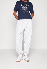 Marineblauw cropped T-shirt met witte tekst "GUESS SPORTS CLUB". Lichtgrijze sweatpants met elastische boorden. Witte sneakers met gumzolen.
