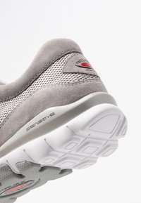 Grå mocka och mesh sneakers med en böjd design, som har en röd accentlogga och en texturerad vit yttersula med ett hexagonalt mönster.