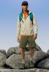 Veste beige légère, chemise orange, shorts verts et sac à dos teal. Pieds dans des chaussures bleues et vertes avec des semelles texturées, debout sur des rochers.