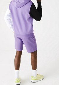 Felpa cappuccio viola chiaro e pantaloni corti coordinati, con una manica nera. Indossati con calze bianche e scarpe da ginnastica giallo pastello.