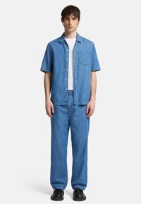 Camicia in denim azzurro chiaro a maniche corte con tasca frontale, abbinata a pantaloni in denim azzurro chiaro a vestibilità comoda e scarpe nere.