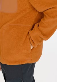 Veste en polaire orange avec une texture douce, dotée d'une poche poitrine zippée et de poches sur les côtés. Pantalon noir partiellement visible.