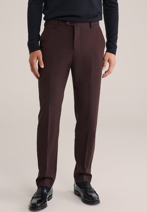 Hombre vestido con pantalones ajustados de color marrón oscuro, mocasines negros y una camiseta de manga larga de color azul marino oscuro, de pie contra un fondo claro y uniforme.