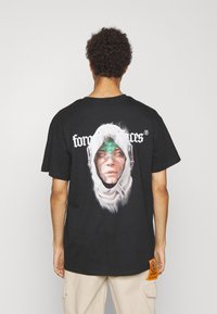 Sort bomulds T-shirt med et stort grafisk motiv af et ansigt i en hætte, grønne accenter og et orange label i kanten med branddetaljer.