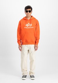 Sudadera con capucha naranja y bolsillo delantero, con logotipo blanco. Combinada con pantalones cargo beiges y zapatillas en blanco y negro.