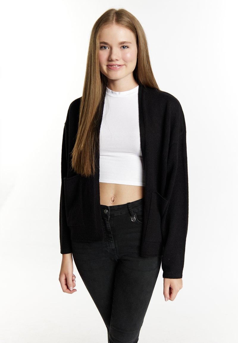 Fekete kardigán struktúrált anyagból, nyitott elülső designnal és két elülső zsebbel, egy fehér crop top felett és fekete farmernadrággal viselve.