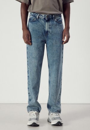 Homme portant un jean bleu délavé droit et des baskets blanches, debout devant un fond uni.