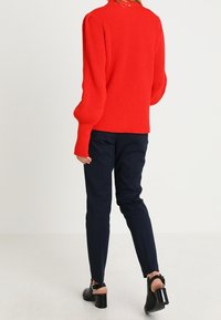 Roter gerippter Pullover mit langen Ärmeln, kombiniert mit navyblauen Slim-Fit-Hosen und schwarzen Schuhen mit Blockabsatz. Dezente Knopfdetails auf der Rückseite.