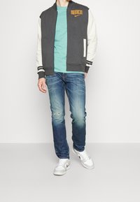Grijze en witte zip-up jas met een Nike-logo, teal t-shirt, blauwe jeans met een vervaagde was, en witte sneakers.