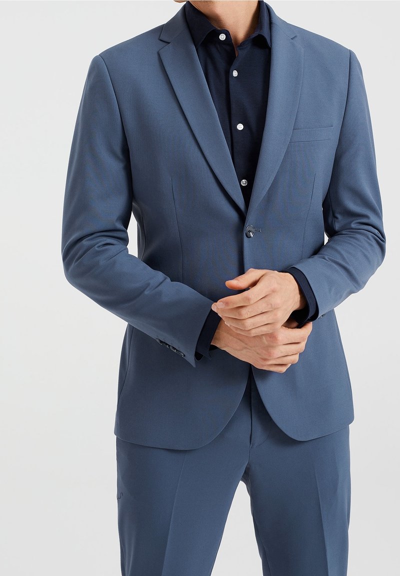 Ljusblå kavaj med notch-lapel, enkel knappstängning och slät textur. Bärs över en marinblå skjorta med krage och knappslå.