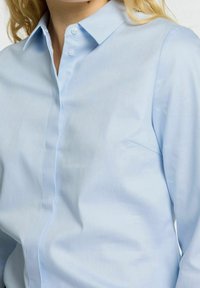 Camicia azzurrina con colletto a punta, realizzata in tessuto liscio, dotata di taschino frontale e patta con bottoni nascosti.