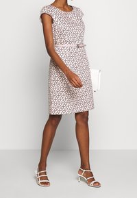 Robe rose avec un motif noir répétitif, manches courtes, silhouette ajustée, associée à des talons blancs à lanières et un petit sac blanc.