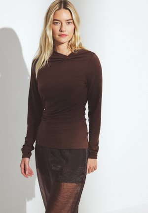 Femme blonde portant un haut brun ajusté à manches longues et une jupe sombre transparente, se tenant contre un fond blanc.